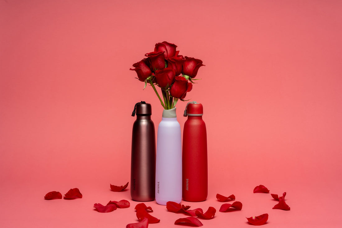 A dark red, a white, and a bright red Avana Ashbury bottle all lined up on a red backdrop with rose bouquet in white bottle. Rose petals scattered around. 